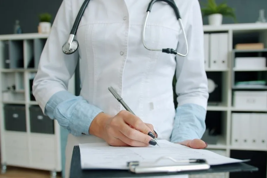 A doctor's hands writing patient notes on a clipboard during a consultation
