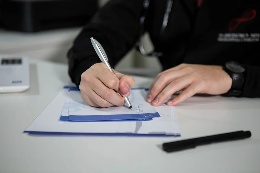 A close-up of a hand writing notes on a clipboard during a clinical consultation